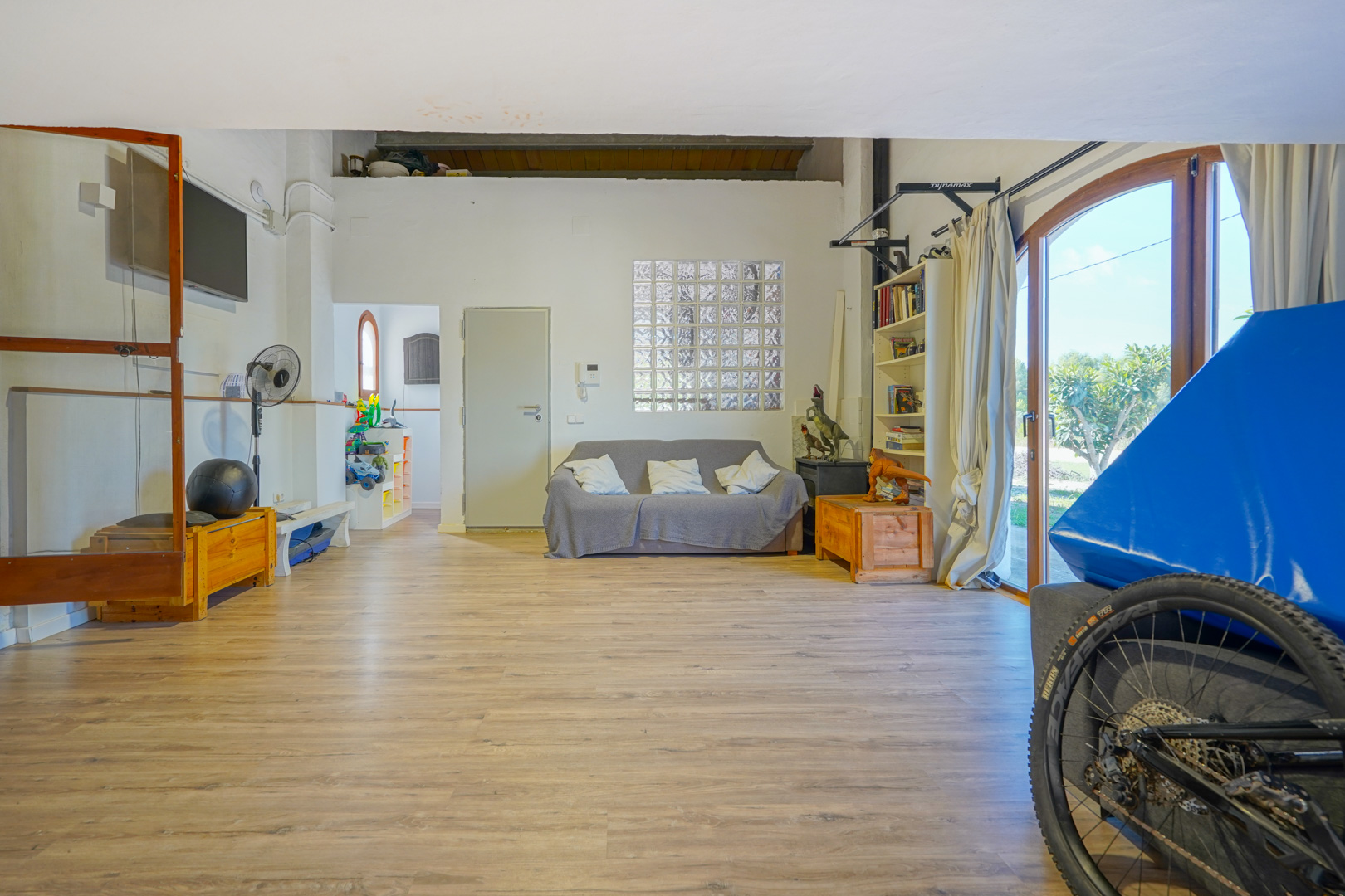 Amplia sala polivalente con suelos laminados, zona de estar y luz natural a través de una ventana de bloques de vidrio y grandes puertas acristaladas.