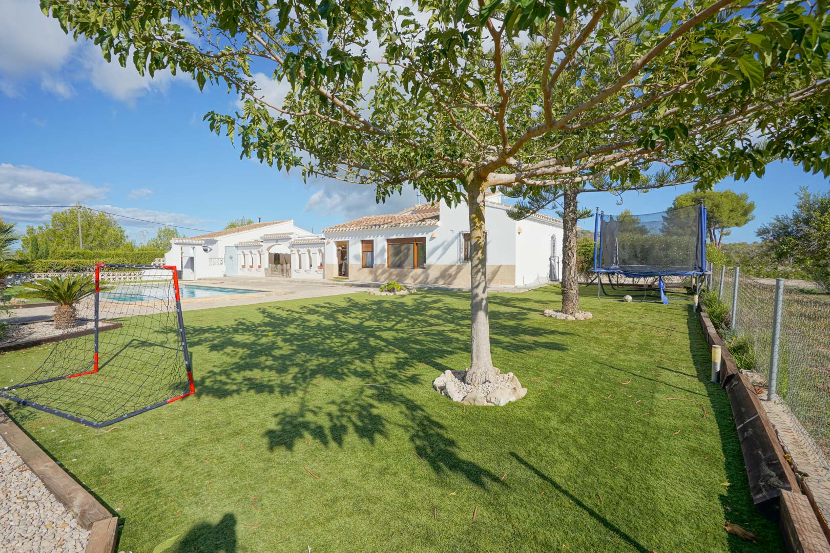 Amplio jardín con césped artificial y zona recreativa que incluye una portería y cama elástica, situado frente a la fachada blanca de la villa y la zona de piscina.