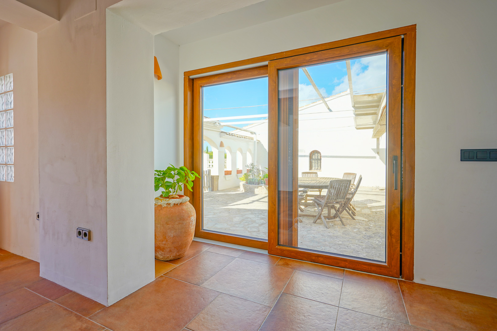 Amplio ventanal corredero con marco de madera que conecta el interior de suelos de terracota con un patio exterior empedrado y zona de comedor al aire libre.