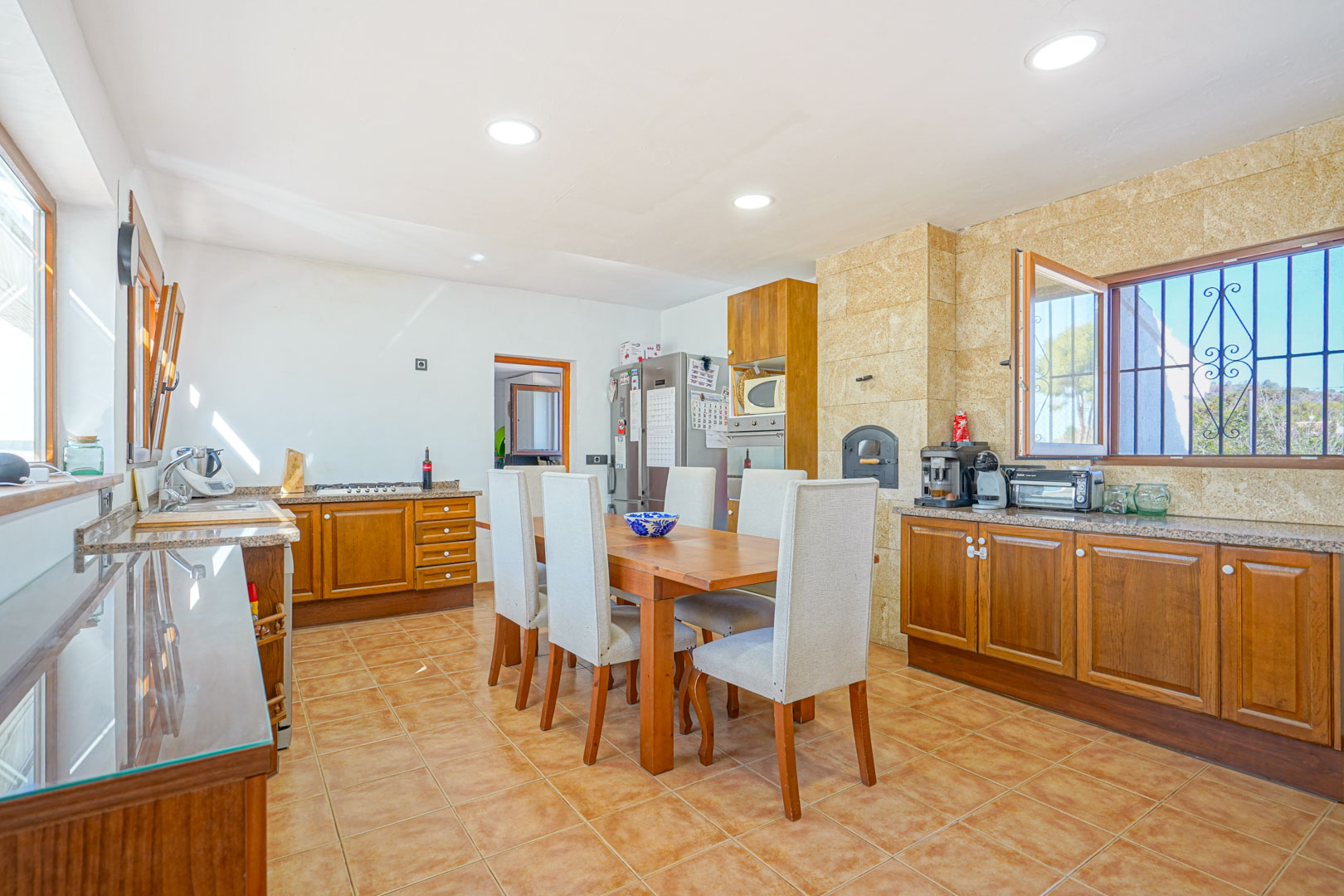 Cocina rústica con armarios de madera, encimeras de granito y un horno de leña integrado en pared de piedra. Incluye zona de comedor central y suelos cerámicos.