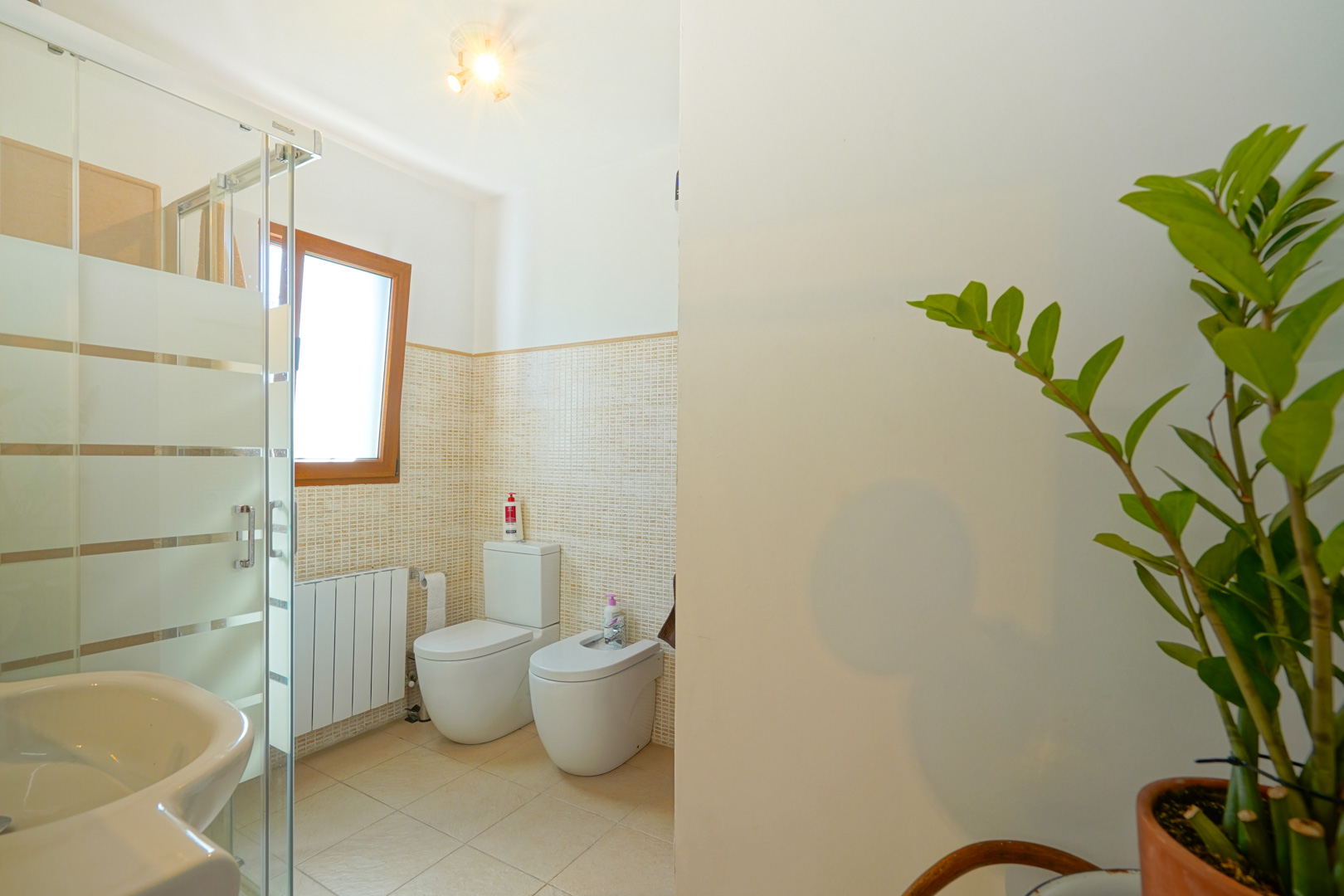 Cuarto de baño con ducha acristalada, inodoro, bidé y radiador, revestido con azulejos de mosaico beige y una ventana con marco de madera.