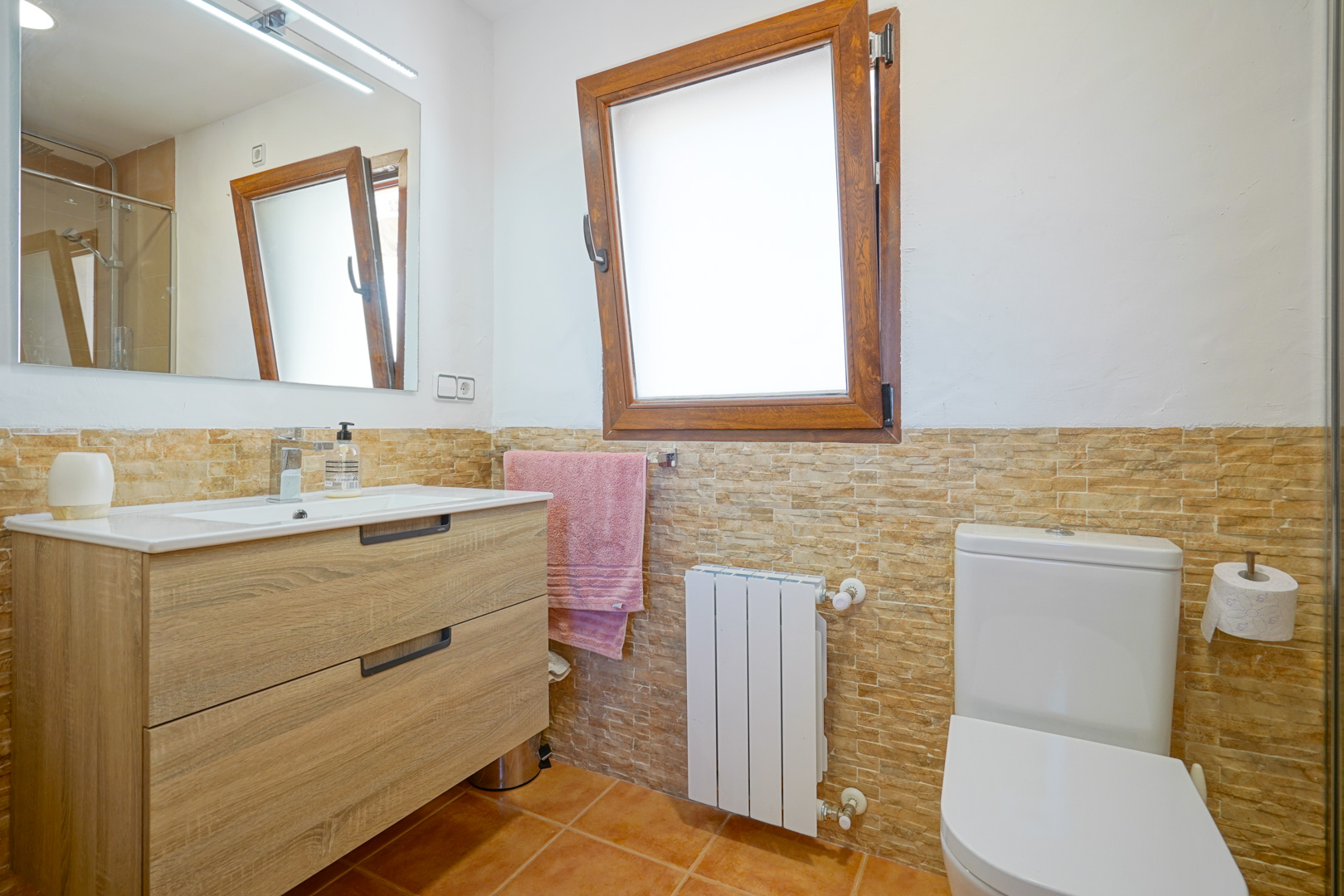Cuarto de baño con mueble de madera, lavabo integrado y espejo con iluminación LED. Incluye paredes con efecto piedra, radiador y ventana oscilobatiente de madera.