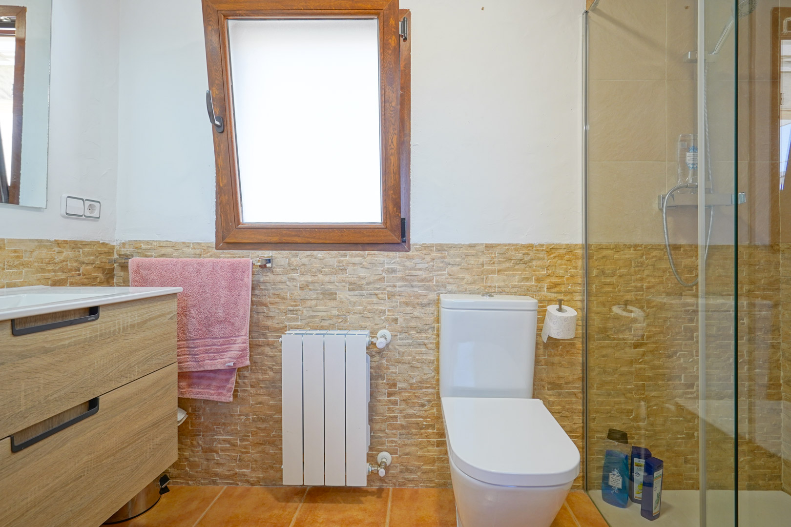 Cuarto de baño con mueble de madera, paredes revestidas de piedra, radiador blanco e inodoro moderno junto a una ducha con mampara de cristal y ventana de madera.