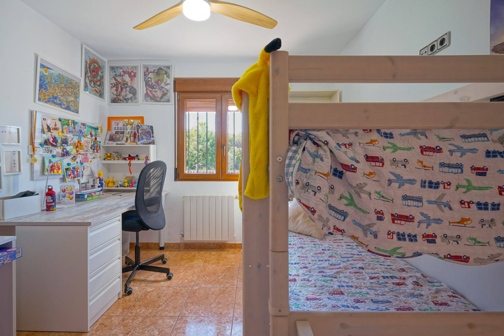 Dormitorio infantil con literas de madera, zona de estudio con escritorio blanco y ventana con marco de madera. Incluye radiador, ventilador de techo y suelo cerámico.