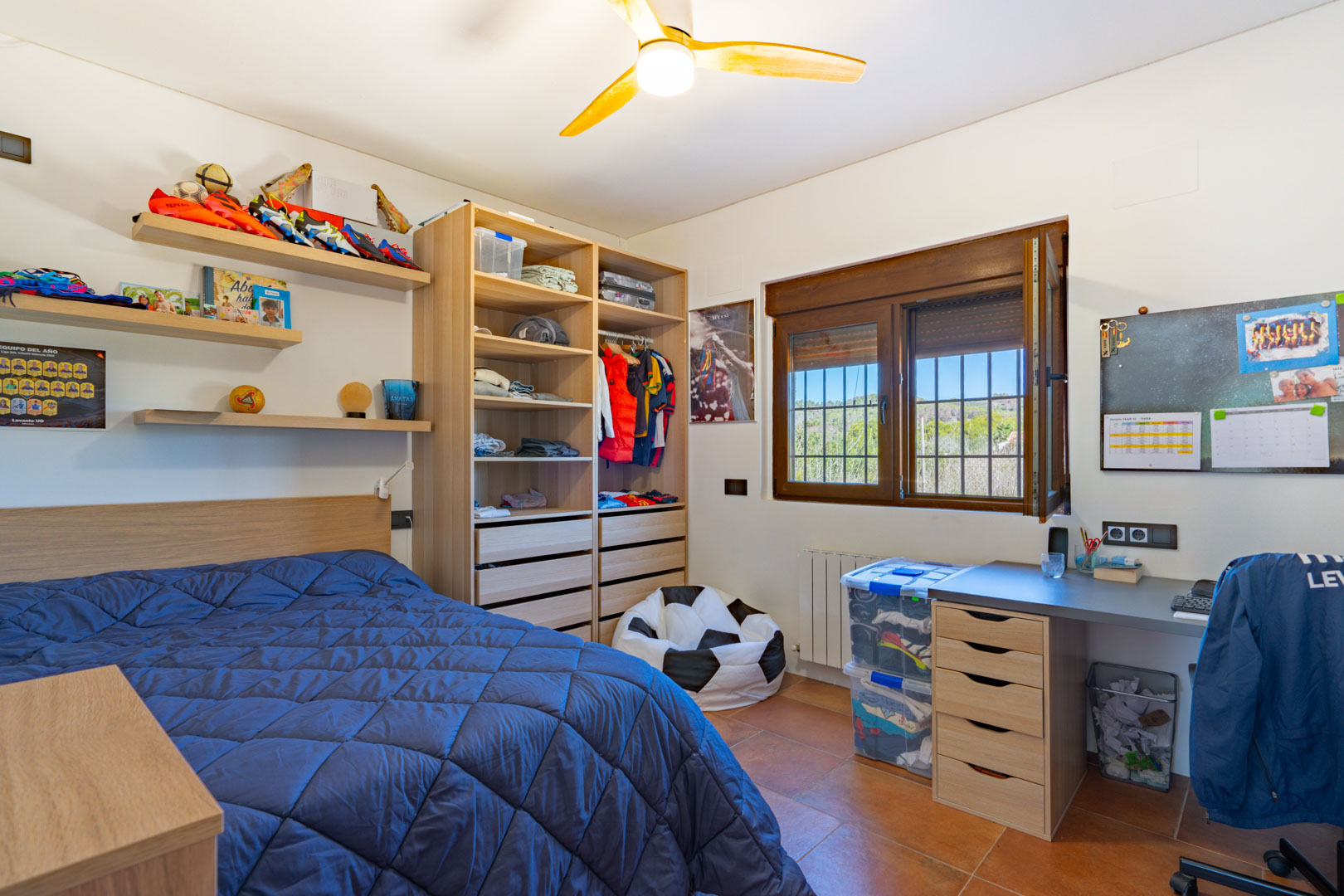 Dormitorio luminoso con suelos de terracota, zona de estudio integrada y armario abierto de madera. Incluye calefacción por radiador y vistas al paisaje natural a través del ventanal.