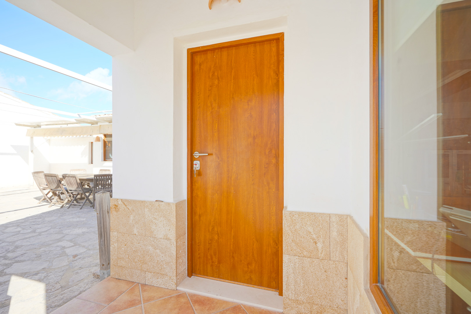 Entrada principal con puerta de madera, zócalo de piedra natural y acceso directo a la terraza exterior pavimentada con mobiliario de madera.