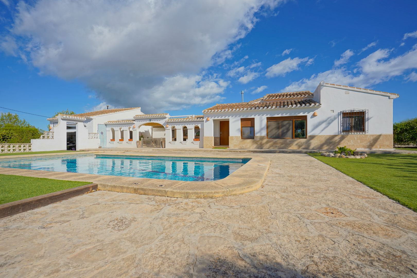 Exterior de la villa con piscina privada, rodeada de una amplia terraza de piedra natural y zonas de césped bajo un cielo despejado.
