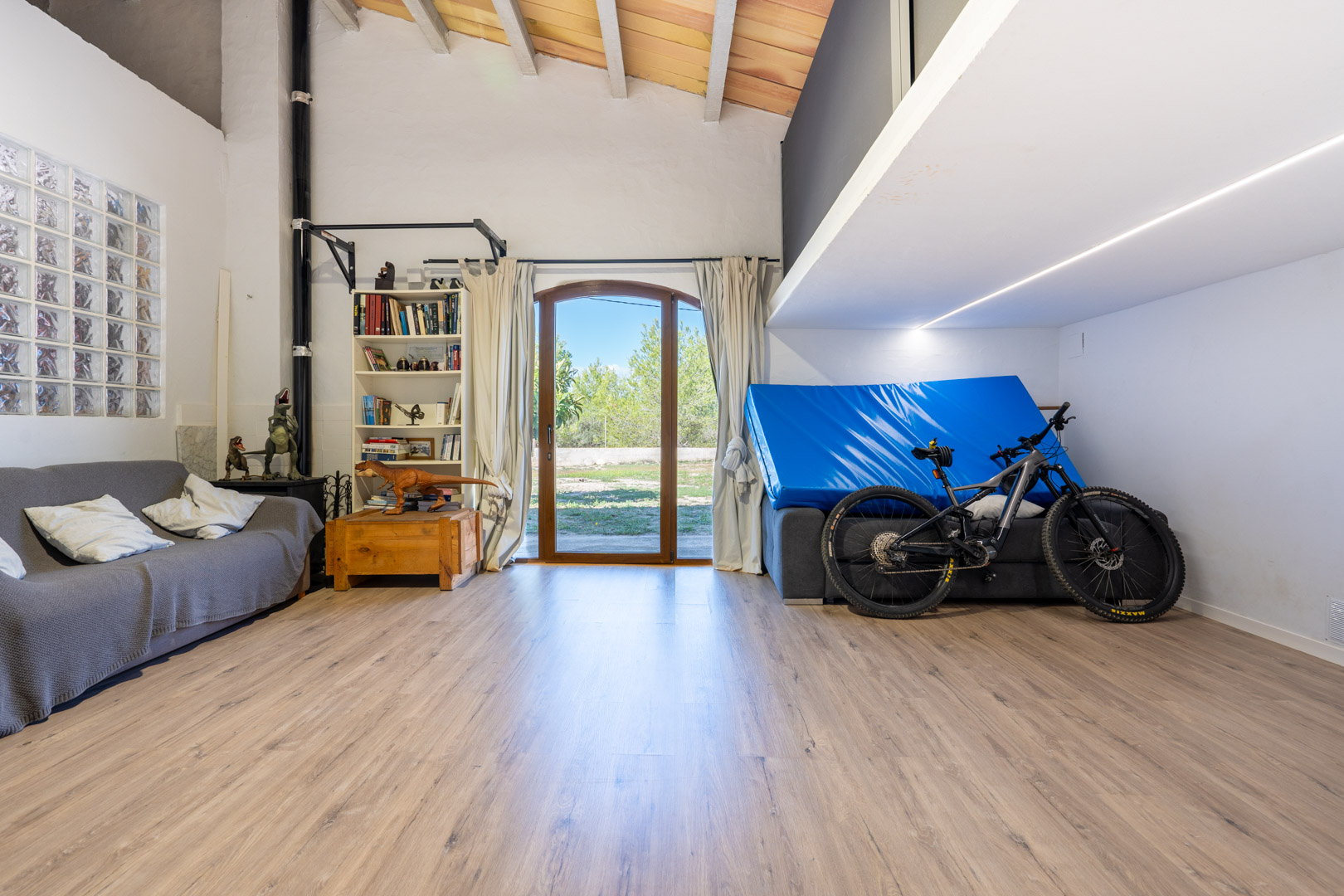 Sala polivalente de techos altos con vigas de madera, suelos laminados, zona de altillo y acceso directo al exterior mediante una puerta acristalada de arco.