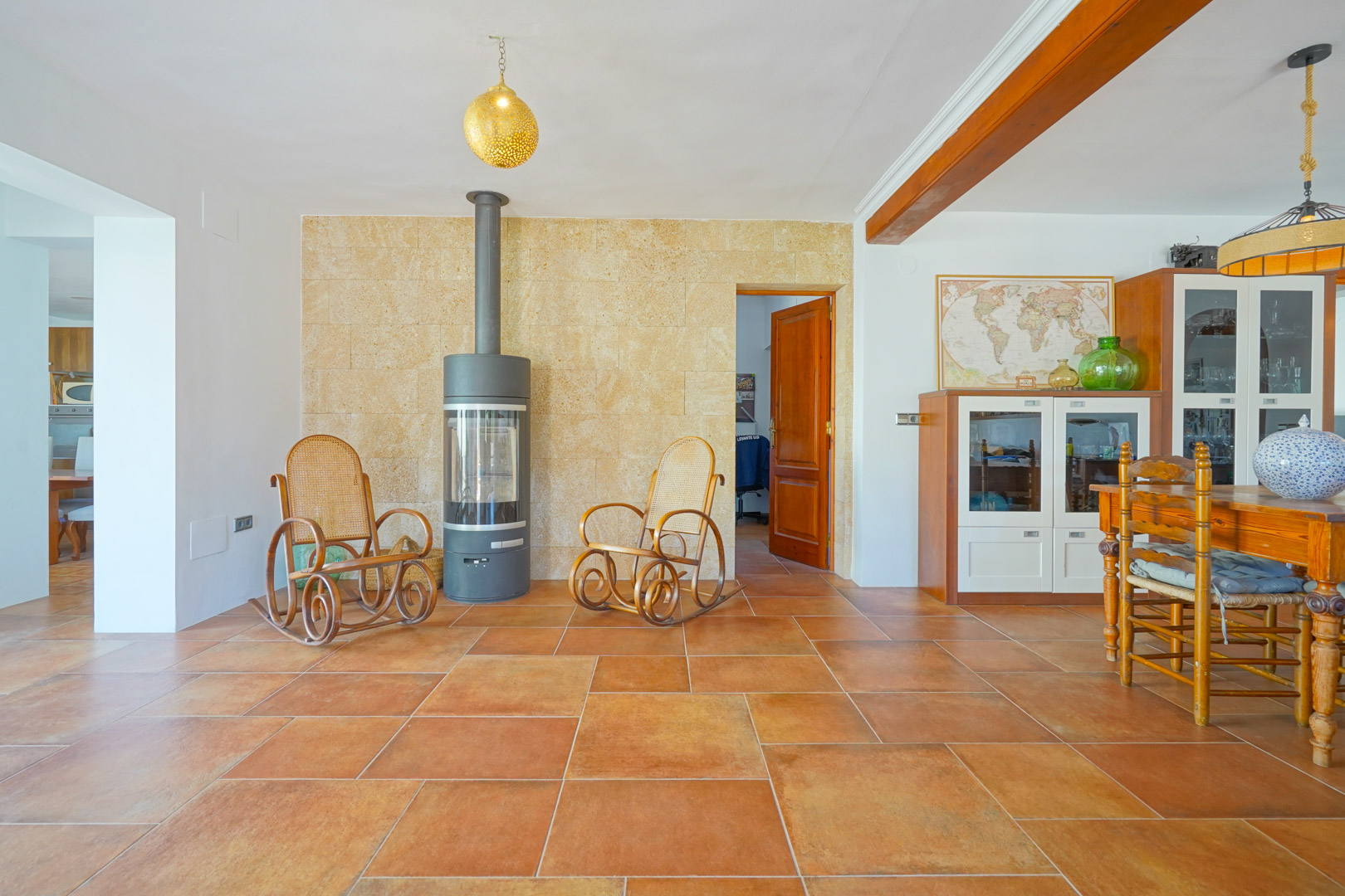 Salón comedor con suelos de terracota, estufa de leña moderna sobre pared de piedra natural y vigas de madera a la vista que aportan un carácter rústico.