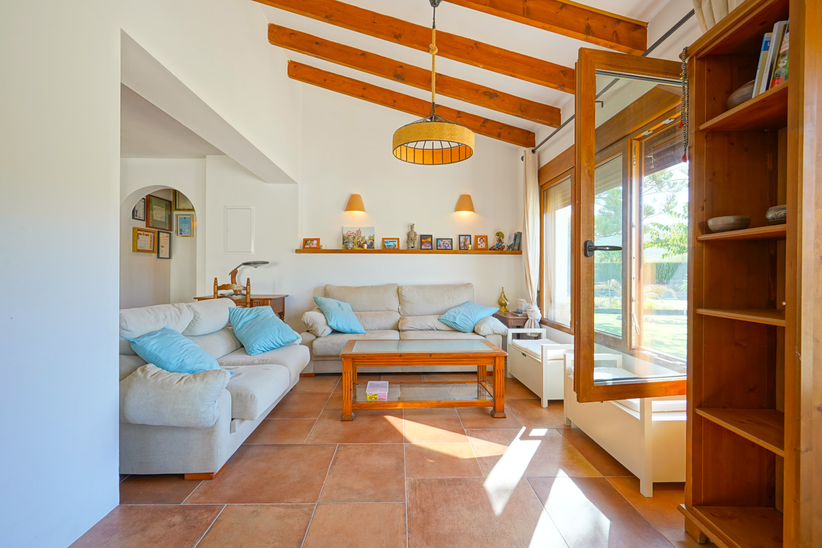 Salón luminoso con techos altos de vigas de madera vistas, suelos de gres rústico y acceso directo al exterior a través de grandes ventanales de madera.
