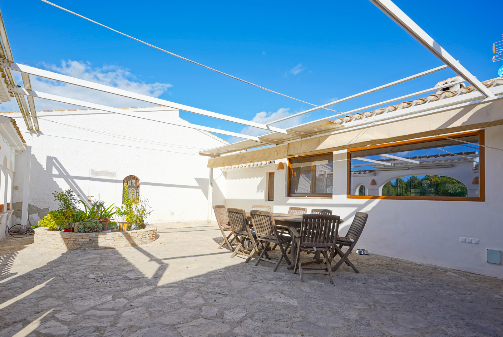 Terraza exterior con suelo de piedra natural, conjunto de comedor de madera y estructura de pérgola metálica junto a una jardinera circular.