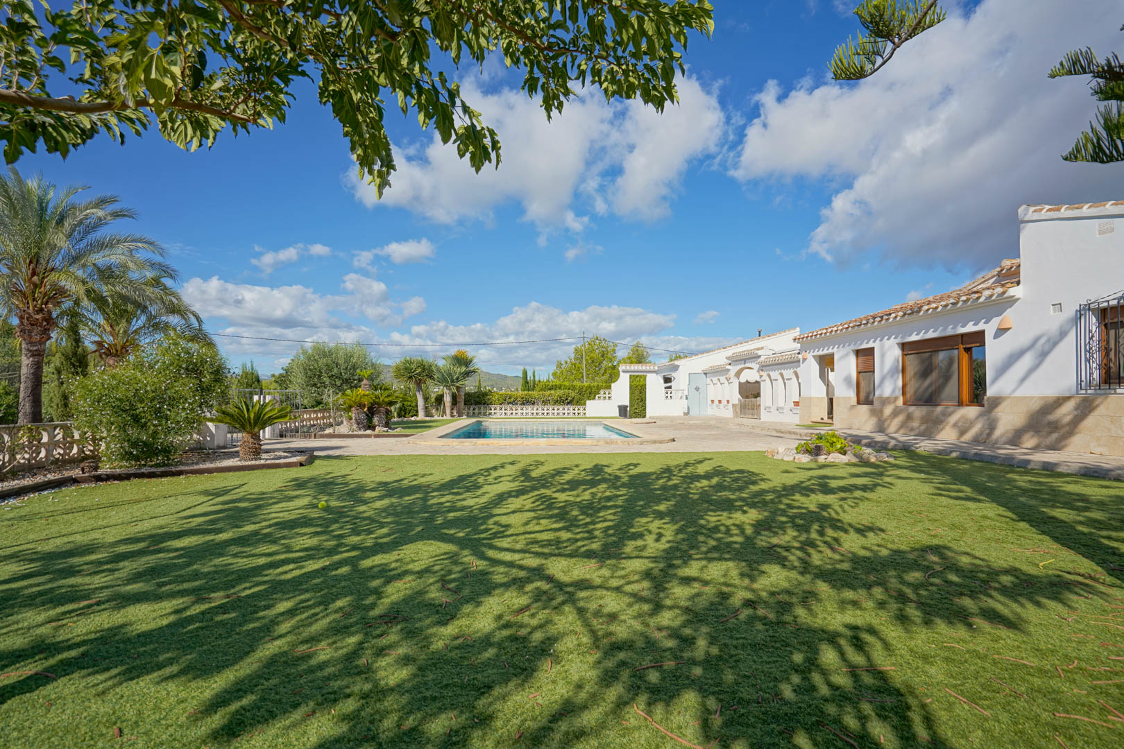 Vista exterior de la villa con fachada blanca y zócalo de piedra, destacando la piscina rectangular rodeada de una terraza soleada y un amplio jardín con césped.