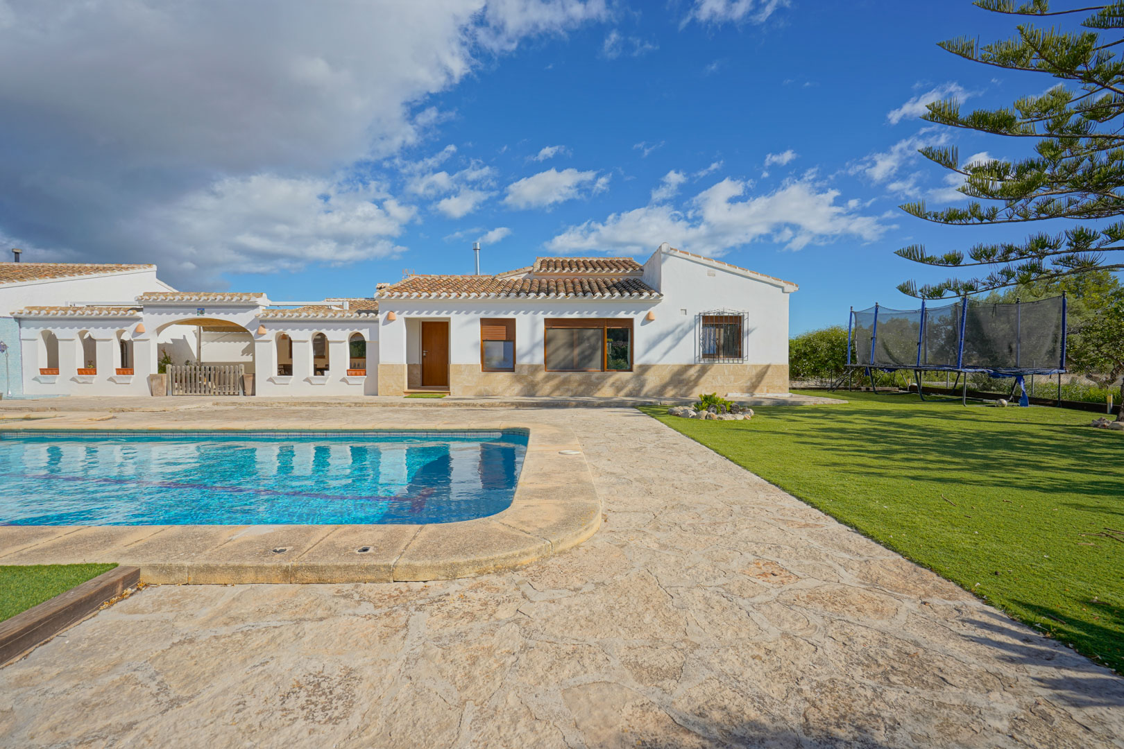 Vista exterior de la villa con piscina privada, amplia terraza de piedra natural y zona de césped, destacando la arquitectura mediterránea de paredes blancas.