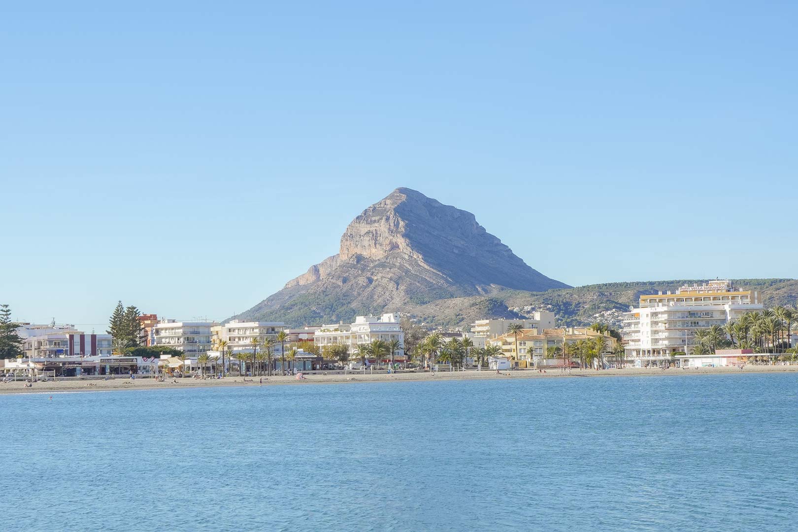 Vista panorámica de la playa del Arenal en Jávea, con el emblemático monte Montgó al fondo y edificios costeros bajo un cielo despejado.