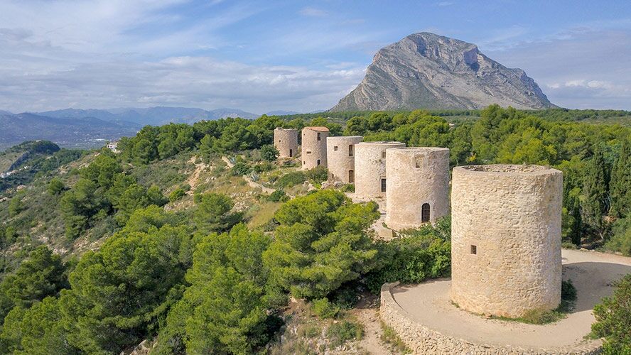 Vista aérea de los emblemáticos molinos de piedra en Jávea, con el imponente macizo del Montgó presidiendo el paisaje natural circundante.