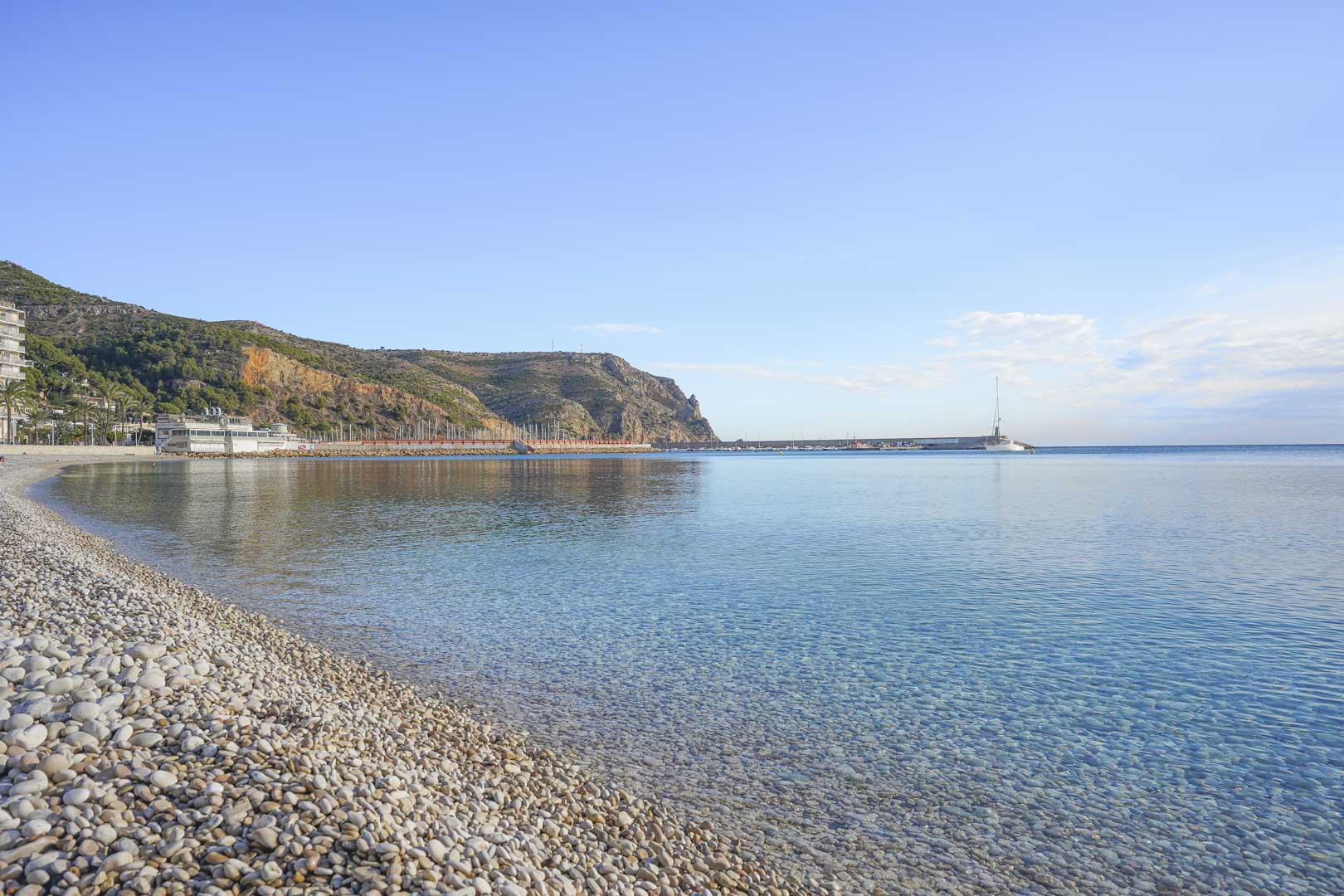 Vista panorámica de la playa de guijarros y aguas cristalinas en Jávea, con el puerto y los acantilados del Cabo de San Antonio al fondo bajo un cielo despejado.