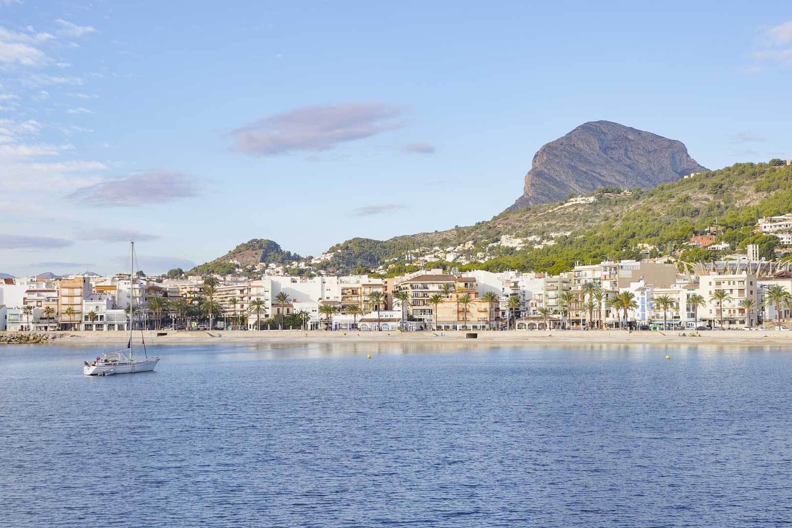 Vista panorámica del puerto y la playa de Jávea con el Montgó al fondo. Un velero navega por las tranquilas aguas frente al paseo marítimo bordeado de palmeras.