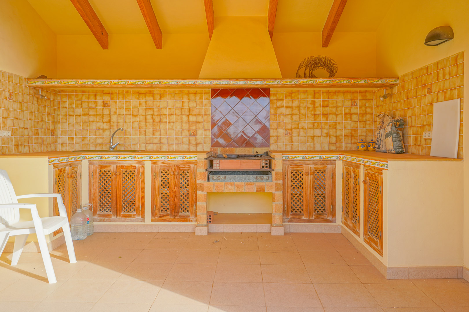 Cocina exterior tradicional con barbacoa de obra, vigas de madera vista y armarios con puertas de celosía bajo un frente de azulejos rústicos.