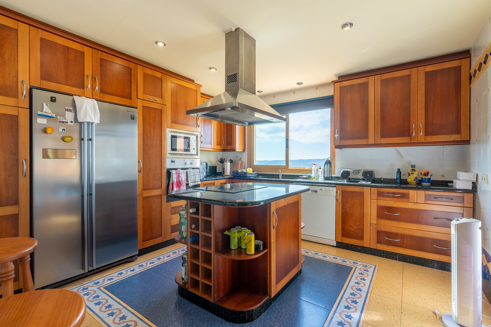 Cocina tradicional con mobiliario de madera, isla central con encimera de granito negro y campana de acero. Dispone de electrodomésticos integrados y vistas al mar desde el ventanal.