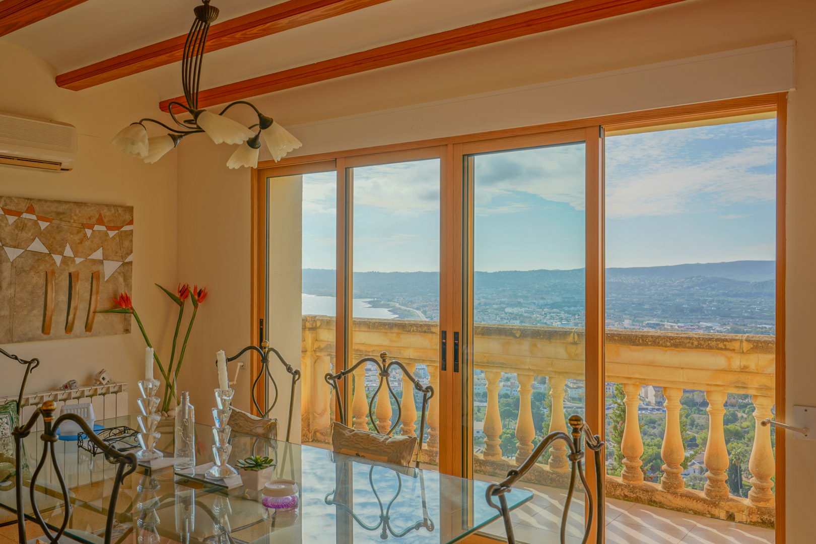 Comedor con mesa de cristal y vigas de madera, con amplios ventanales que ofrecen vistas panorámicas de la bahía de Jávea y la costa mediterránea.