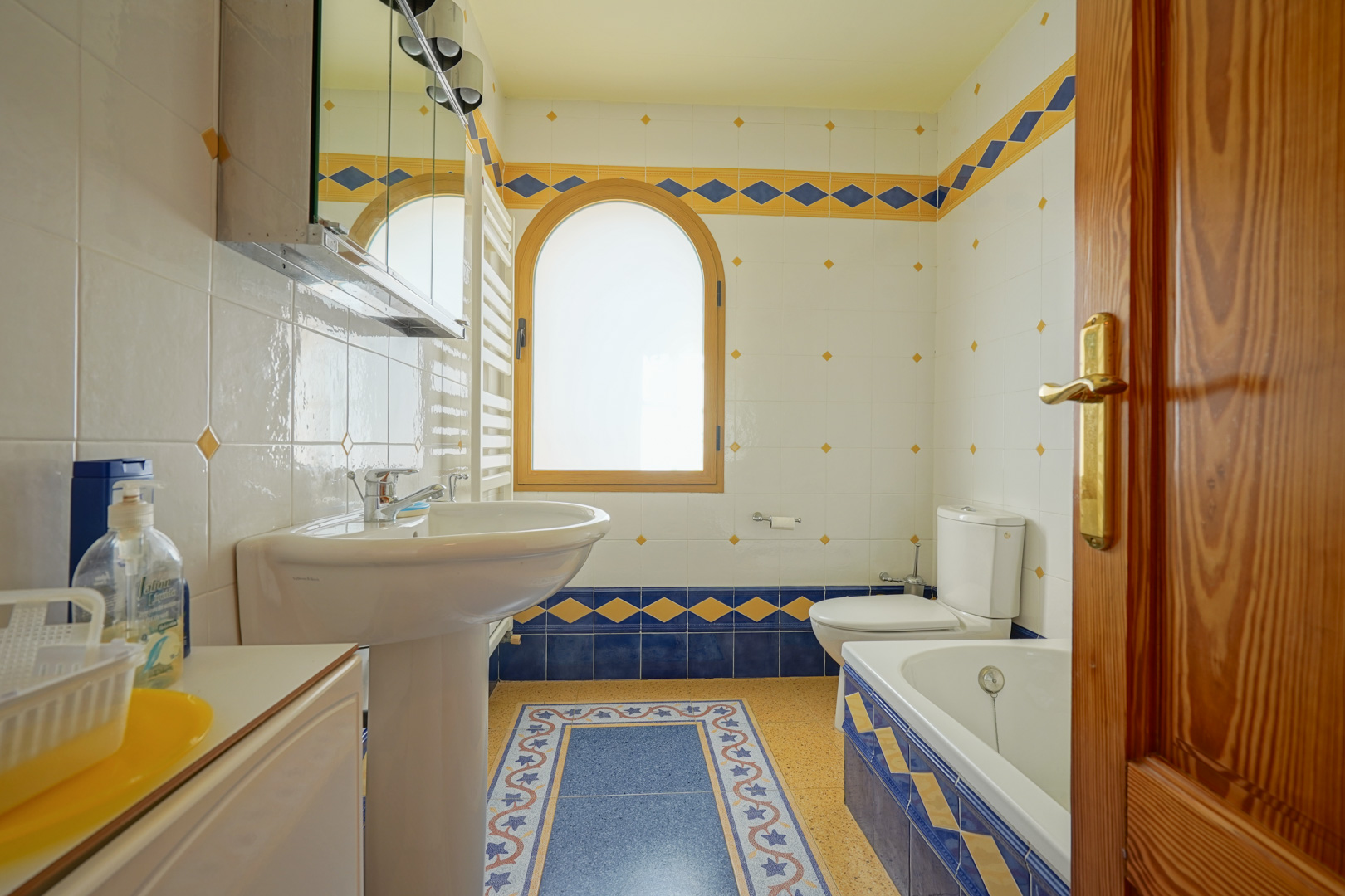 Cuarto de baño con azulejos decorativos en azul y amarillo, ventana de arco con marco de madera y equipamiento funcional que incluye bañera, lavabo de pedestal y radiador toallero.