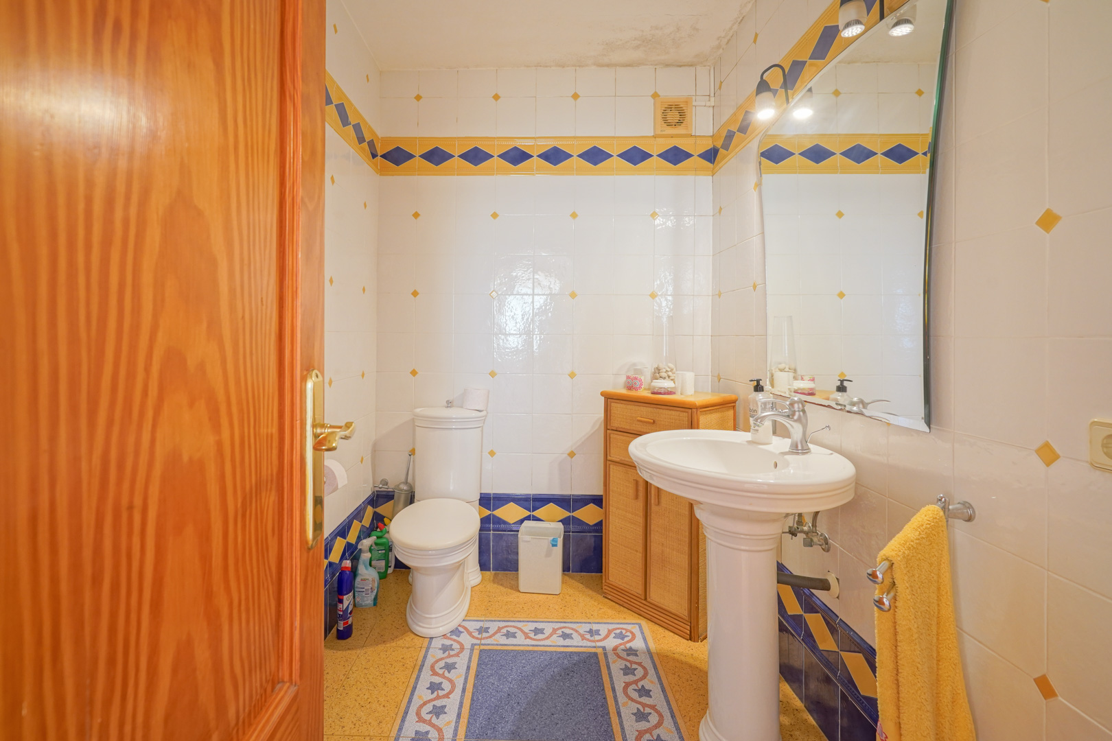 Cuarto de baño de estilo tradicional con azulejos blancos, cenefas en azul y amarillo, lavabo de pedestal y mueble auxiliar de madera.