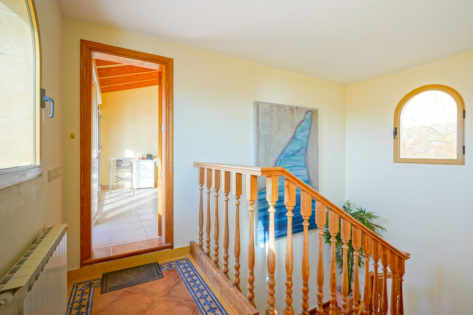 Distribuidor con escalera de madera, suelos de terracota con cenefa decorativa y ventana de arco. El acceso conduce a una estancia luminosa con techos de vigas de madera.