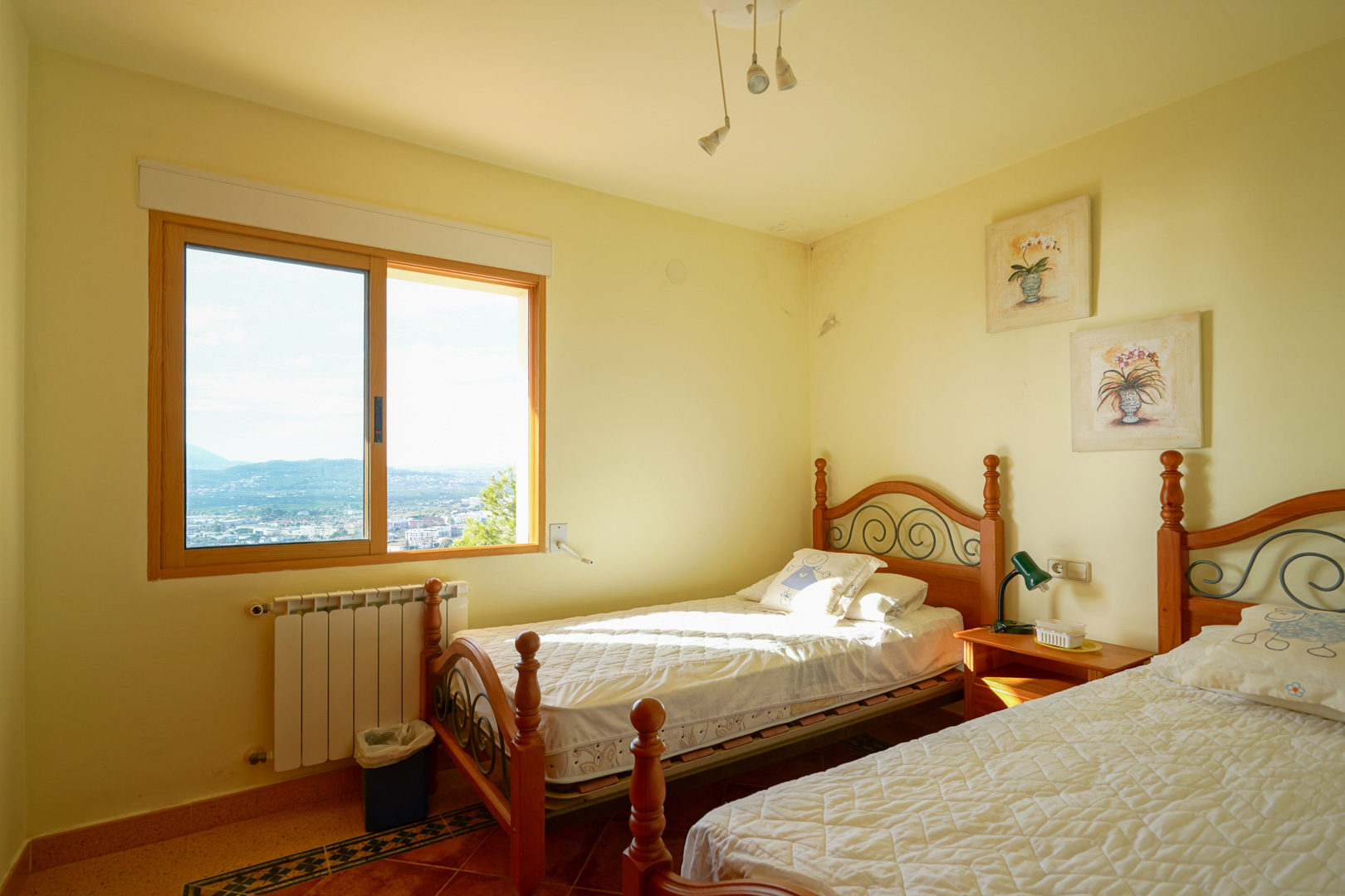 Dormitorio doble con camas de madera y forja, suelos cerámicos y ventana con marco de madera que ofrece vistas despejadas al valle y las montañas.