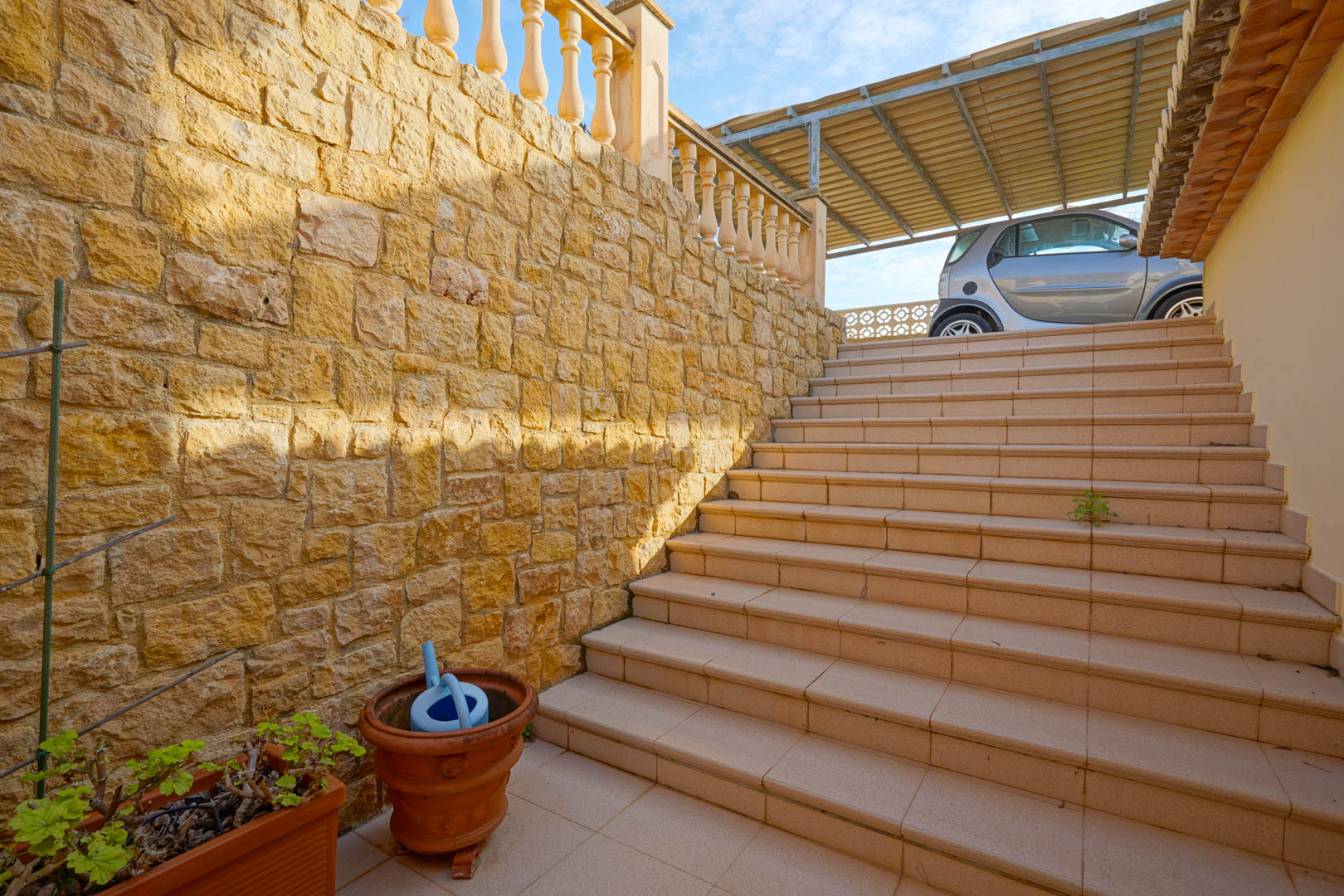 Escalera exterior flanqueada por un muro de piedra natural que conecta la entrada principal con la zona de aparcamiento superior y el porche cubierto.