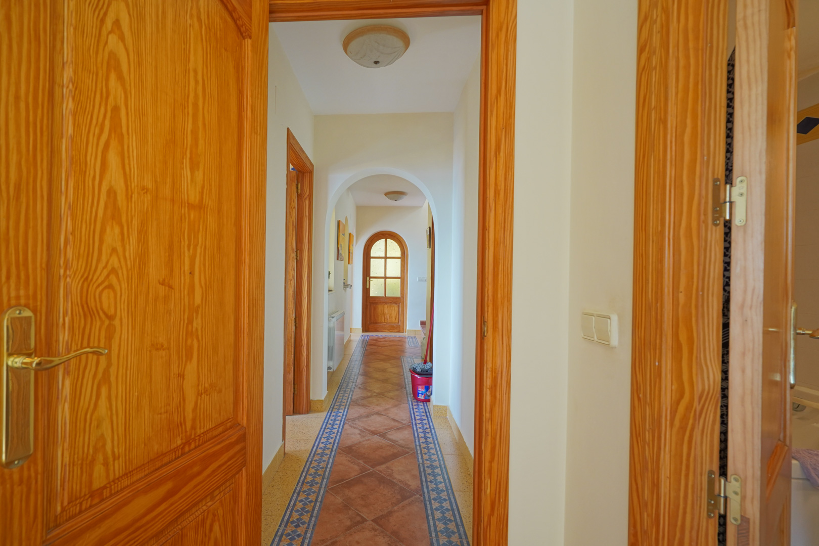 Pasillo con arcos tradicionales, suelos de baldosa cerámica con cenefas decorativas y carpintería de madera natural que conecta las estancias.