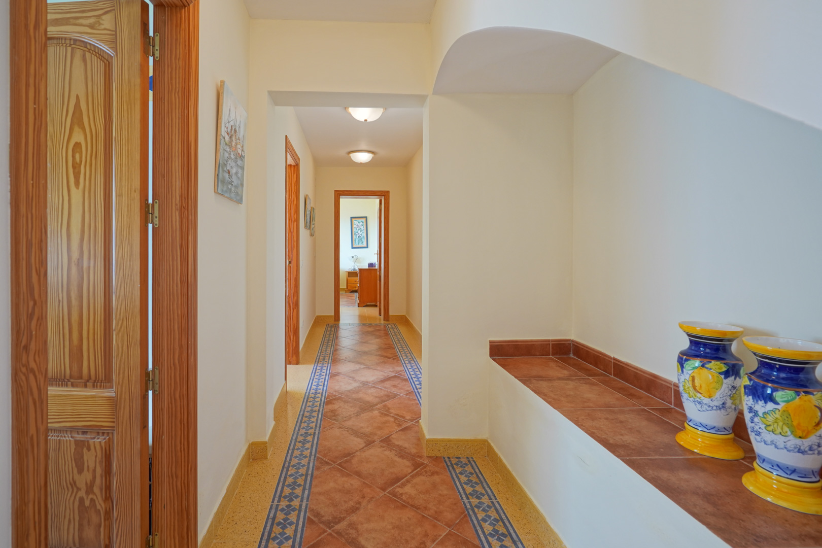 Pasillo con suelos de baldosas cerámicas y cenefas decorativas, puertas de madera natural y un banco de obra integrado con jarrones ornamentales.