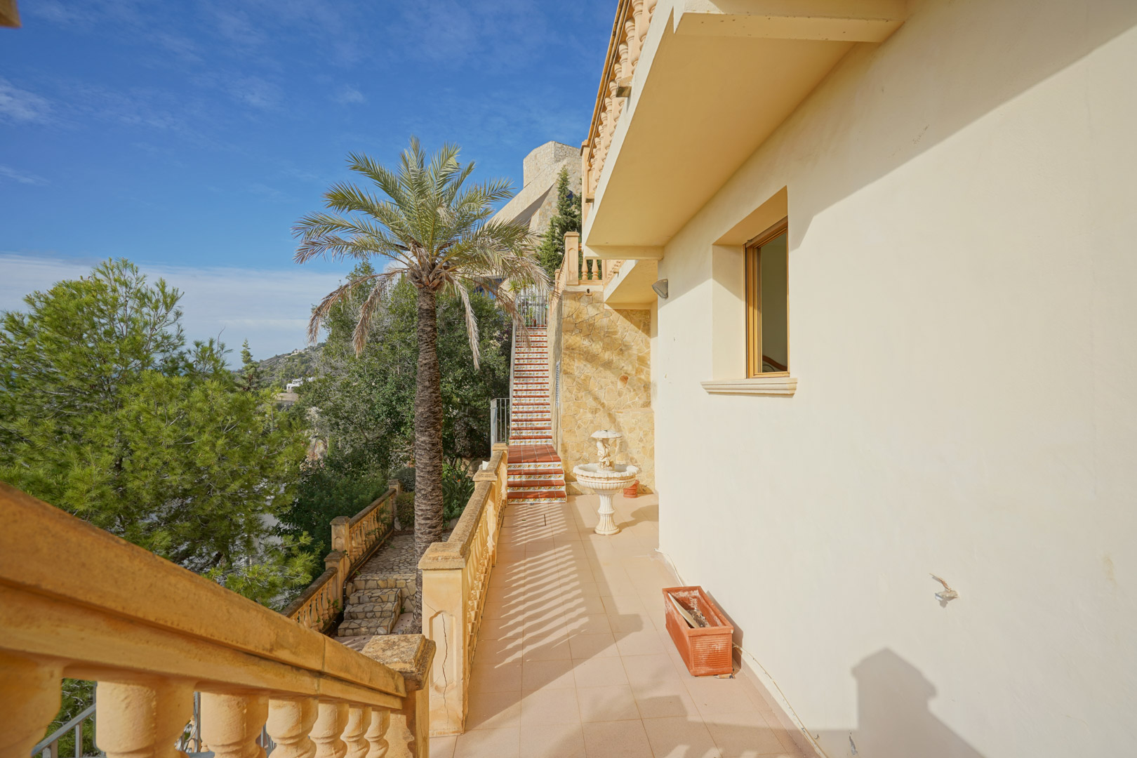 Pasillo exterior con balaustrada clásica, muros de piedra natural y escalera decorada con azulejos tradicionales junto a una palmera.
