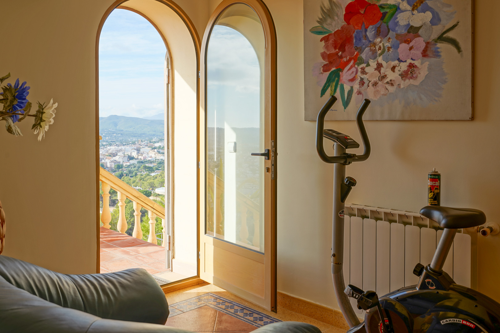 Sala interior con bicicleta estática y puertas de arco que abren hacia una terraza con vistas panorámicas al valle y las montañas de Jávea.