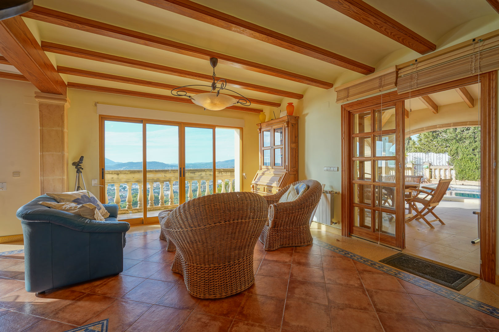 Salón con vigas de madera vista, suelos de terracota y mobiliario de mimbre. Los ventanales ofrecen vistas panorámicas al valle y acceso directo a la terraza exterior.
