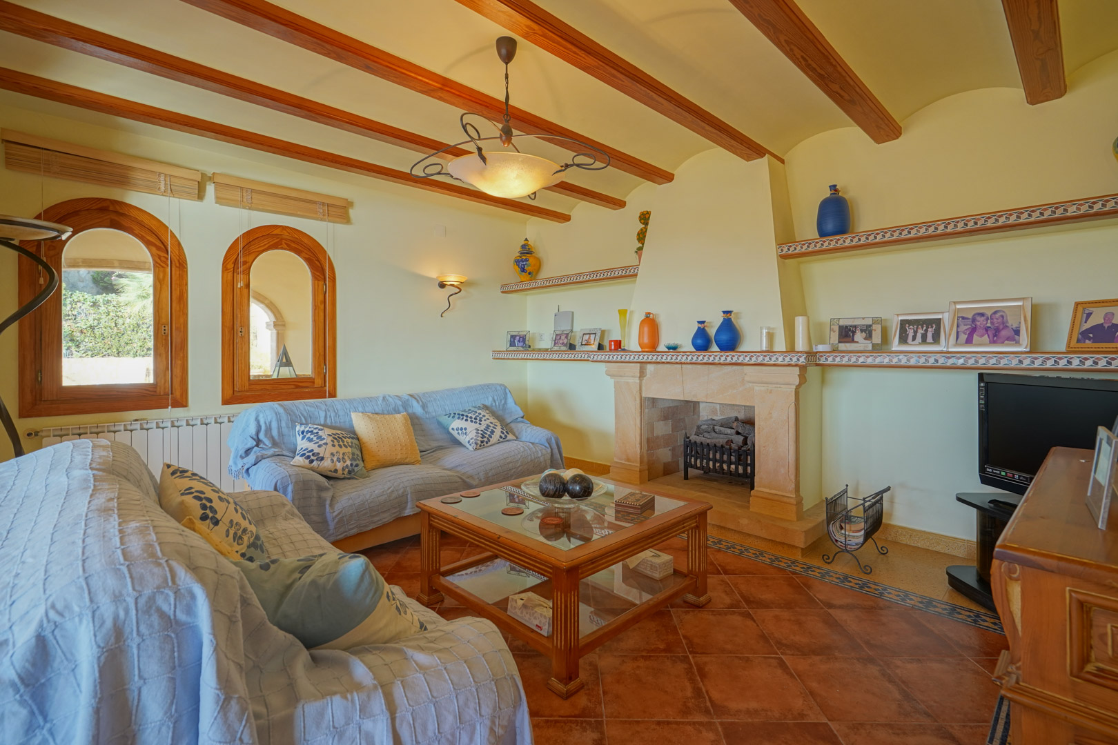 Salón tradicional con chimenea de piedra, vigas de madera vista y suelo de terracota. Incluye ventanas de arco de madera y estanterías integradas con detalles cerámicos.