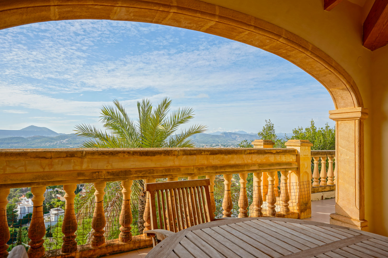 Terraza cubierta con arco de piedra tradicional y balaustrada, ofreciendo vistas panorámicas a las montañas y al valle de Jávea con mobiliario de madera.