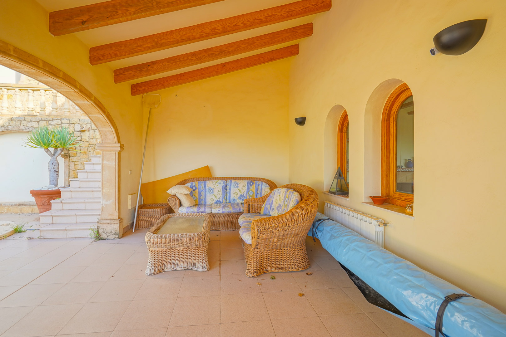 Terraza cubierta con vigas de madera a la vista, mobiliario de mimbre y arcos tradicionales que conectan con el exterior y la zona de piscina.