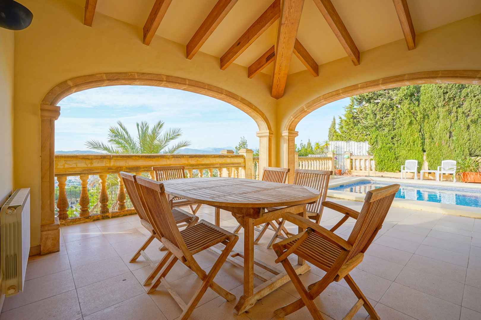 Terraza cubierta con vigas de madera y arcos de piedra, equipada con comedor exterior frente a la piscina y vistas panorámicas a la montaña.