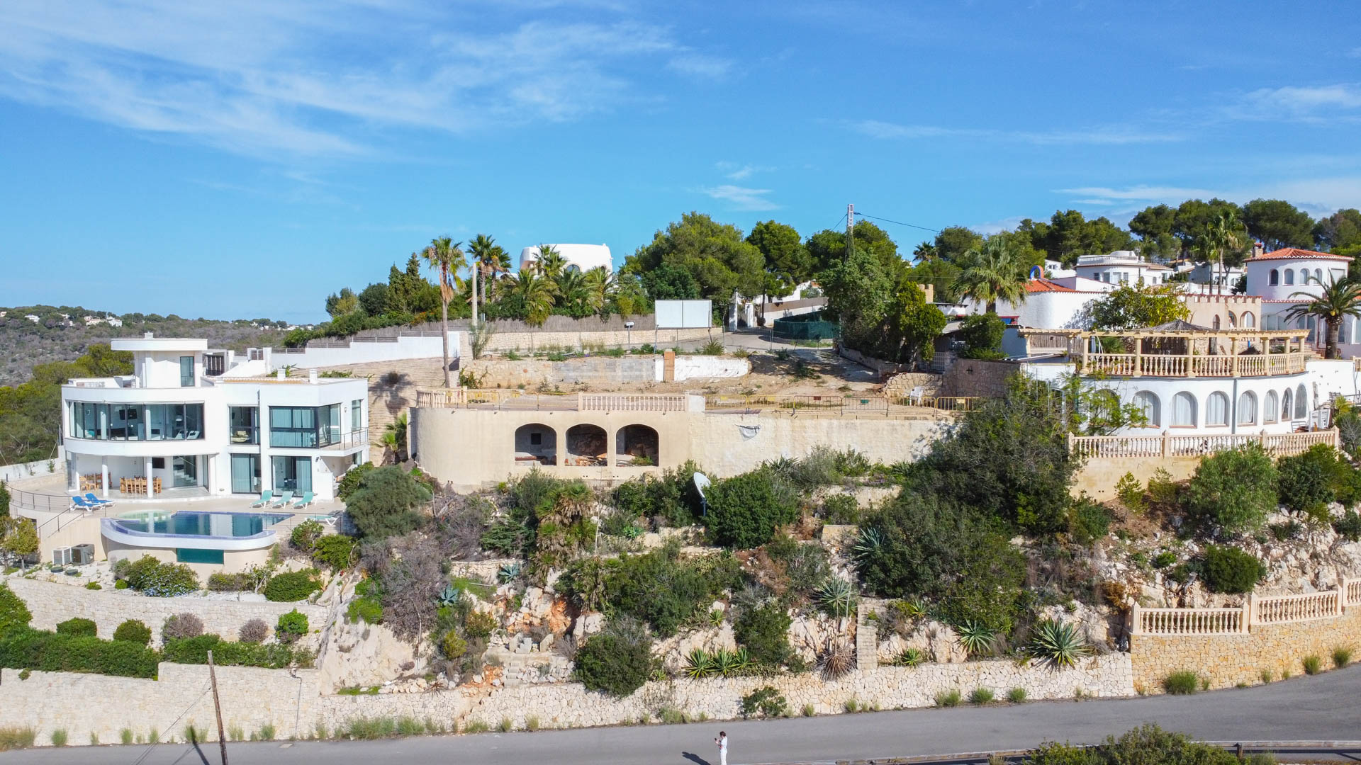 Vista aérea de la parcela elevada con estructura de piedra arqueada, rodeada de villas de lujo y vegetación mediterránea autóctona sobre muros de contención.