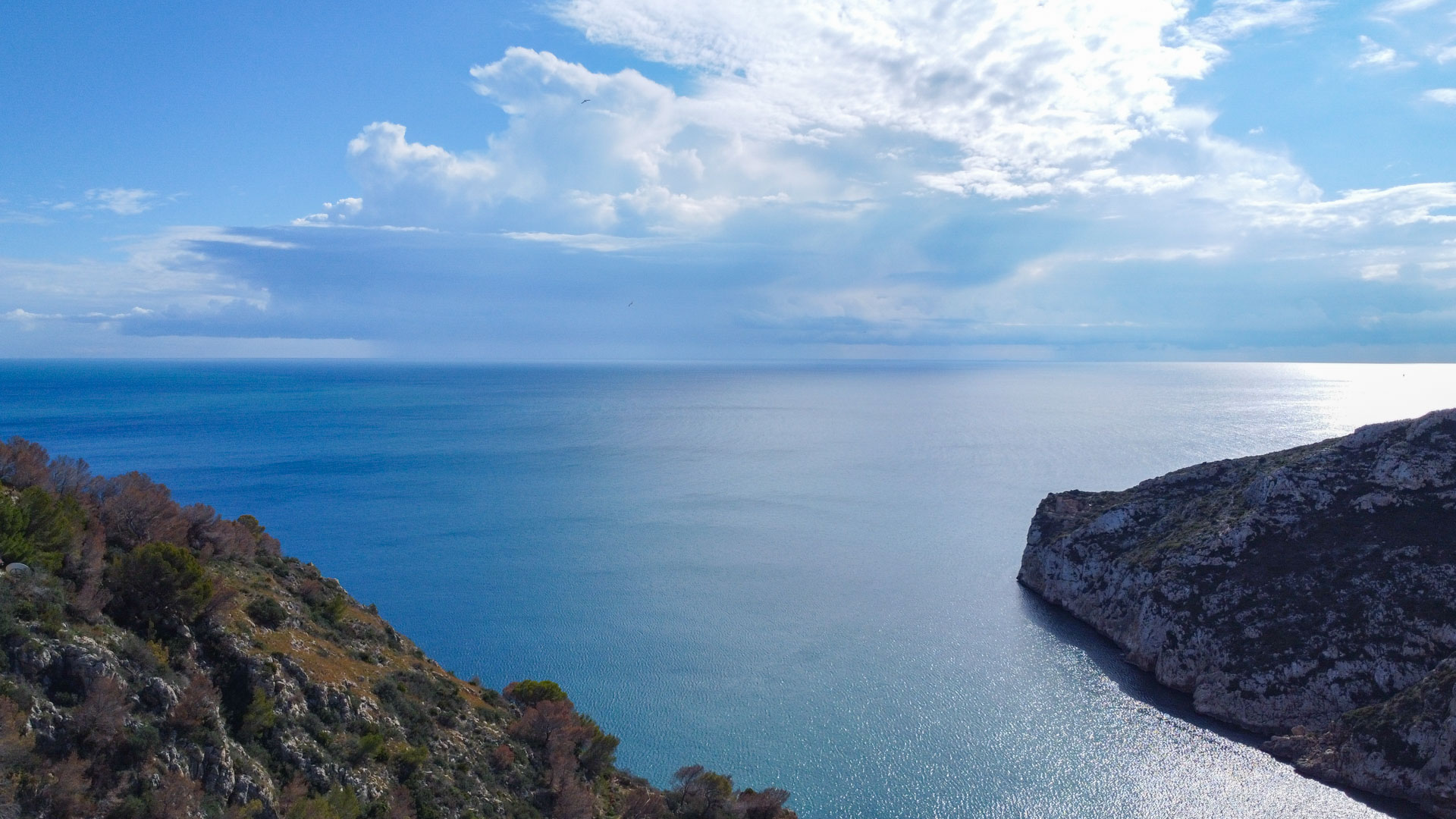 Vista aérea frontal del mar Mediterráneo y los acantilados rocosos que flanquean la bahía de la Granadella bajo un cielo parcialmente nublado.