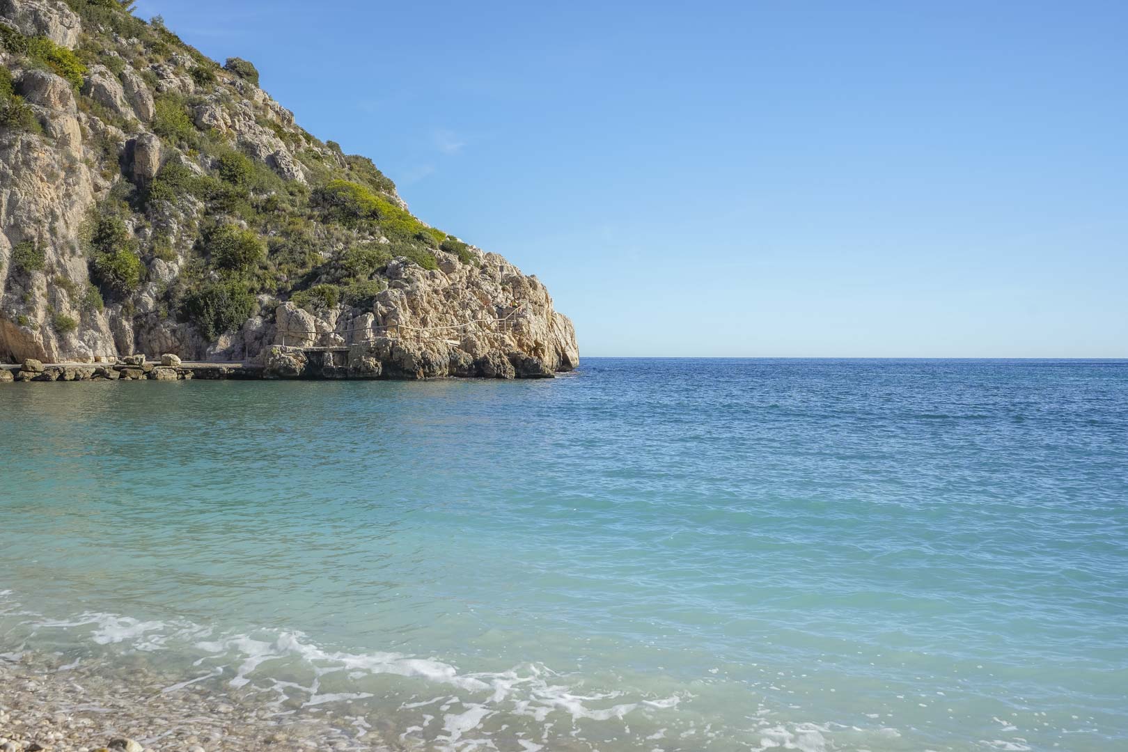 Vista de las aguas cristalinas color turquesa de la Cala Granadella, rodeada de acantilados calcáreos y vegetación mediterránea bajo un cielo despejado.