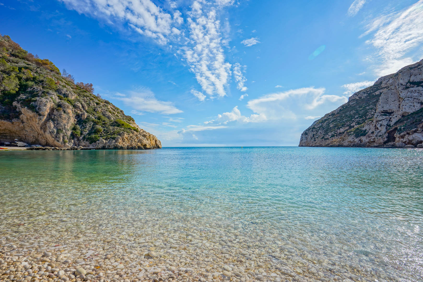 Vista panorámica de la playa de la Granadella con sus características aguas cristalinas y orilla de cantos rodados, flanqueada por acantilados escarpados bajo un cielo despejado.