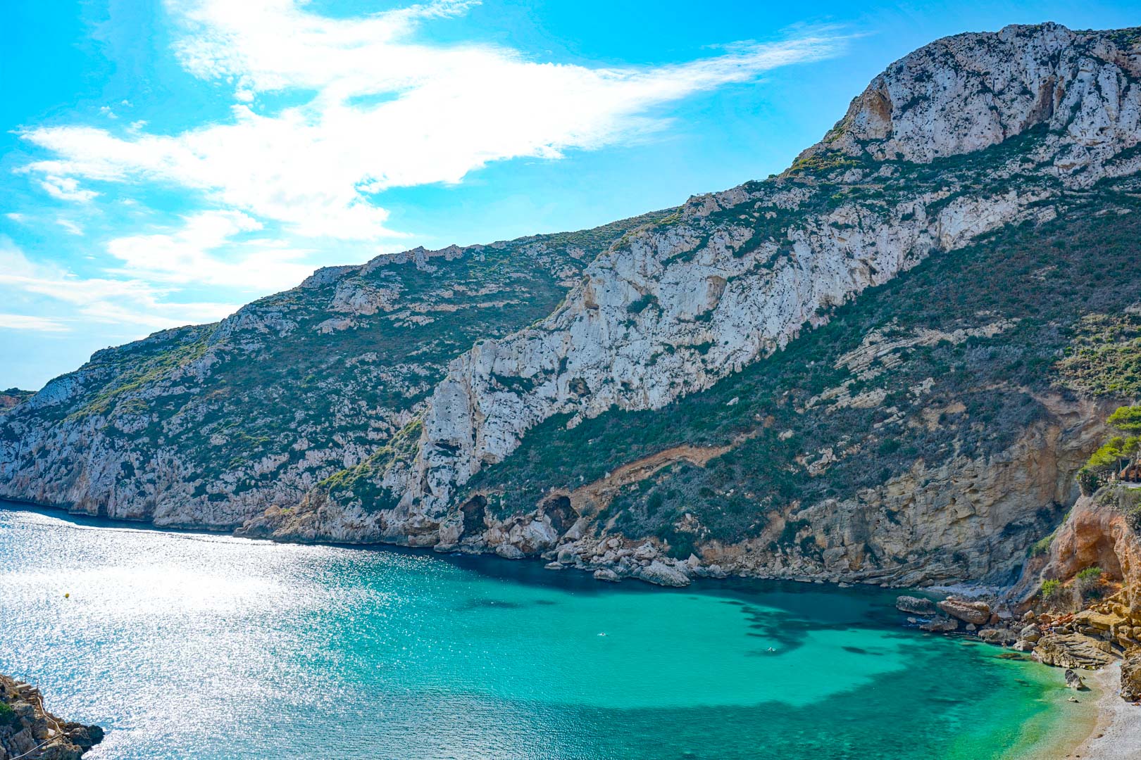 Vista panorámica de las aguas turquesas de la Cala Granadella, rodeada de acantilados calizos y vegetación mediterránea bajo un cielo despejado.