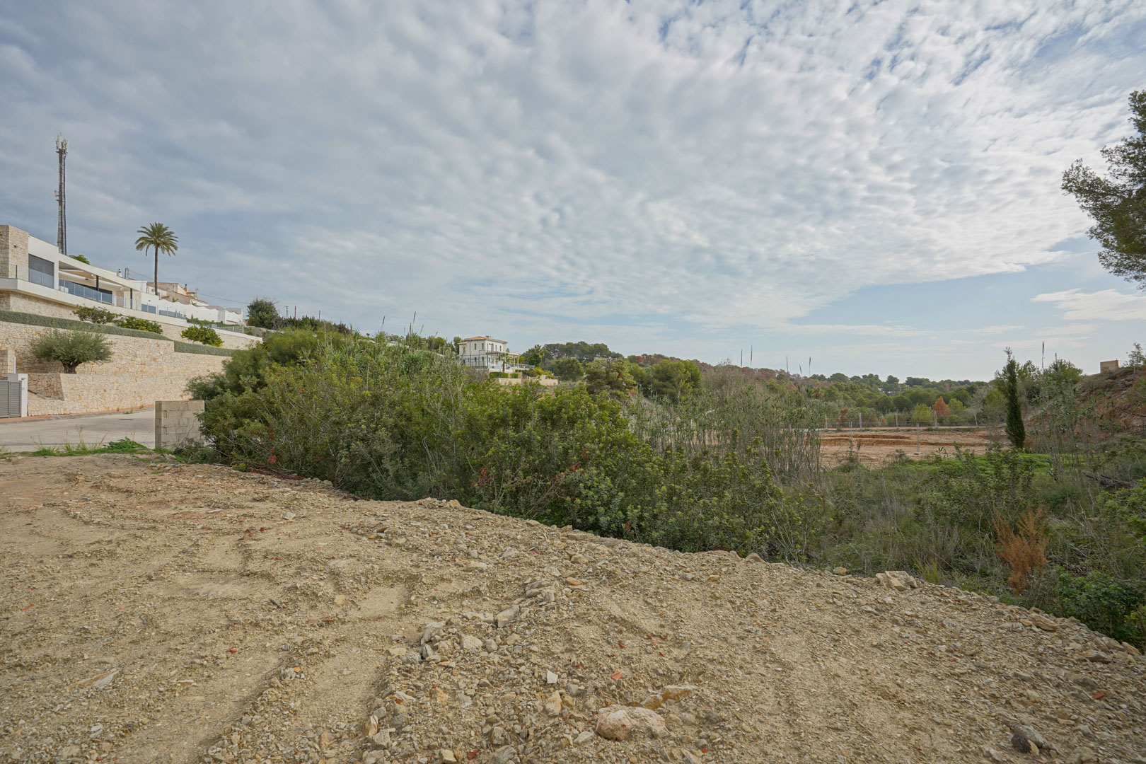 Parcela de terreno preparada para la construcción, rodeada de vegetación mediterránea y villas modernas con muros de piedra natural en una zona residencial consolidada.