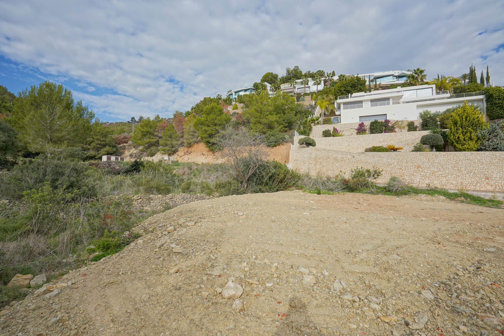 Parcela elevada en preparación rodeada de pinos, con vistas a villas modernas de arquitectura blanca y muros de piedra seca integrados en la ladera.