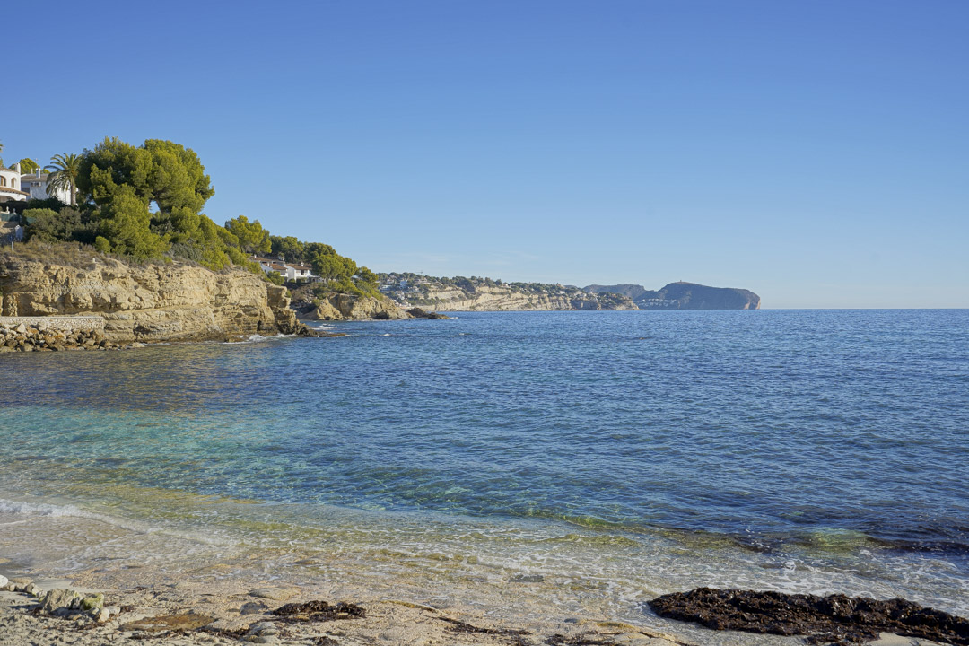 Vista de una cala de aguas cristalinas en Calpe, con acantilados de piedra caliza y pinos mediterráneos bajo un cielo despejado.