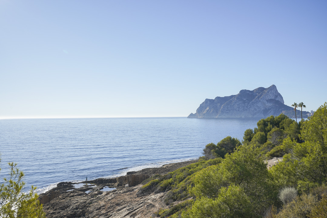 Vista panorámica del Peñón de Ifach y la costa mediterránea desde Calpe, destacando la vegetación autóctona y el mar azul bajo un cielo despejado.