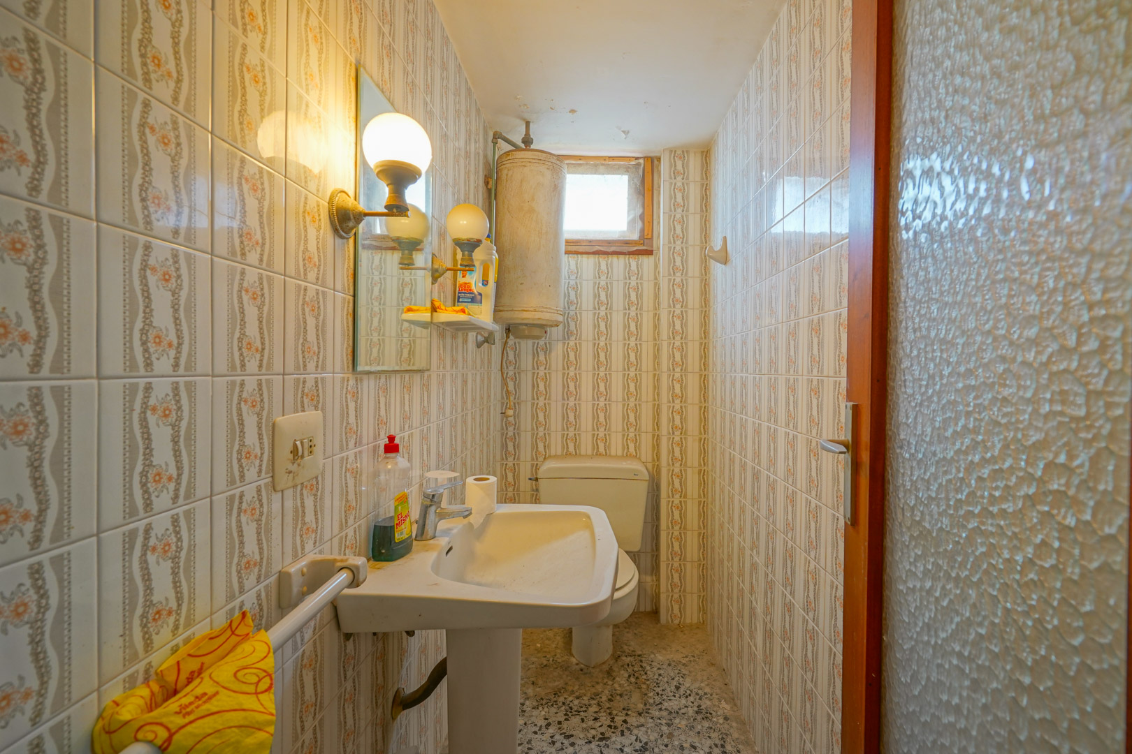 Cuarto de baño funcional con azulejos decorativos de época, lavabo de pedestal, termo eléctrico y suelos de terrazo original.