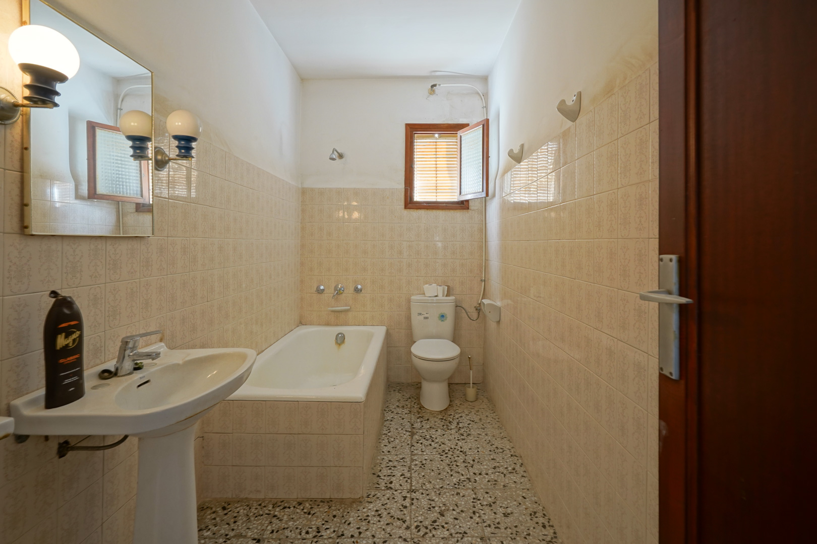 Cuarto de baño funcional con bañera encastrada, lavabo de pedestal, suelos de terrazo y paredes alicatadas con azulejos clásicos.