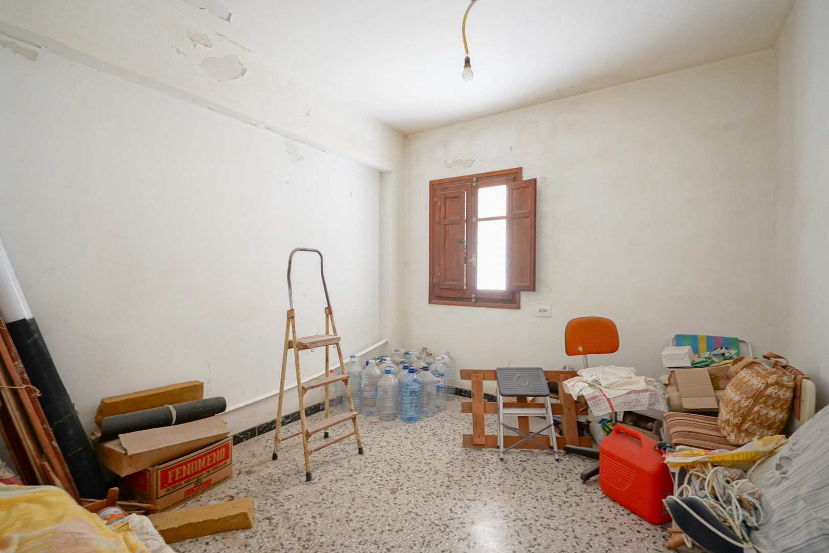 Habitación con suelo de terrazo y ventana de madera con contraventanas. El espacio muestra el estado original y el potencial de reforma integral de la estancia.